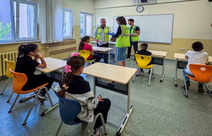 Görselde bir sınıf ortamı görülüyor. Sıralarda oturan çocuklar önde, tahtanın önünde ise yeşil yelek giymiş yetişkinler ayakta duruyor ve öğrencilerle ilgileniyor. Çocuklar dikkatle onları dinlerken, yetişkinler belgeler üzerinde çalışıyor gibi görünüyor. Ortam, eğitim veya bilgilendirme amaçlı bir etkinliği andırıyor.
