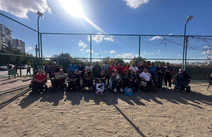 Görselde açık havada, voleybol sahasında bir grup kişi toplu halde poz veriyor. Katılımcıların bir kısmı tekerlekli sandalyede, bazıları ise ayakta duruyor. Arkada tel örgü, ağaçlar ve mavi gökyüzü görünüyor. Güneşli bir günde çekilmiş.