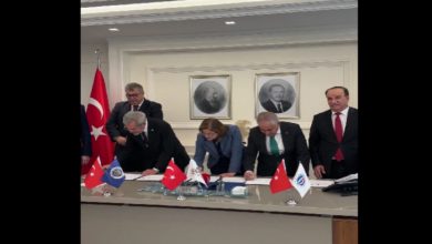 Görselde, bir toplantı salonunda masa etrafında toplanmış kişiler protokol imzalarken görülüyor. Masada Türkiye, Gaziantep Büyükşehir Belediyesi ve üniversitelere ait bayraklar yer alıyor. Arka planda Atatürk portresiyle birlikte başka bir portre asılı.