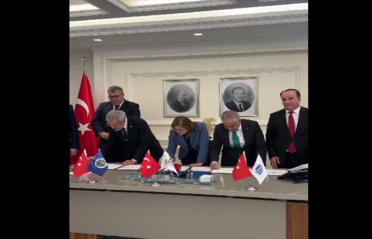 Görselde, bir toplantı salonunda masa etrafında toplanmış kişiler protokol imzalarken görülüyor. Masada Türkiye, Gaziantep Büyükşehir Belediyesi ve üniversitelere ait bayraklar yer alıyor. Arka planda Atatürk portresiyle birlikte başka bir portre asılı.