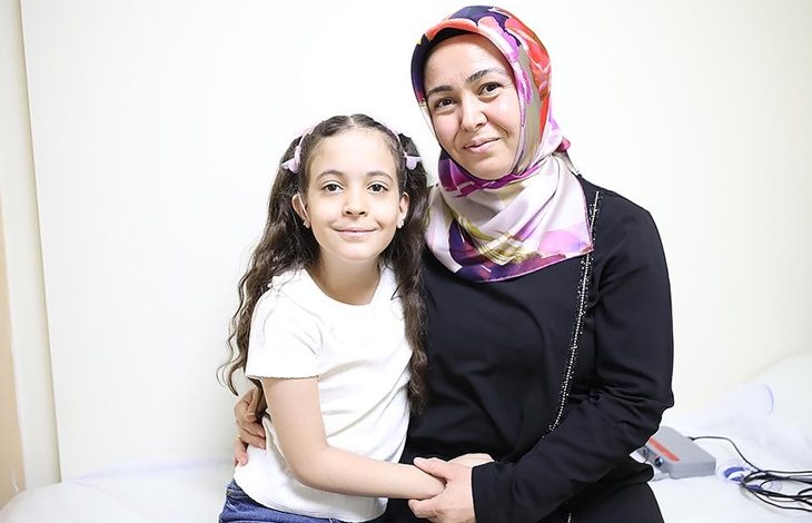 Görselde bir kadın ve küçük kızı yan yana oturmuş, gülümseyerek poz veriyor. Kadın başörtülü, siyah kıyafetli ve kızını kucaklamış. Küçük kız ise beyaz tişört giymiş, saçları iki yandan toplanmış. İkisi de samimi ve mutlu bir şekilde kameraya bakıyor. Ortam sade bir iç mekân, muhtemelen bir hastane odası.