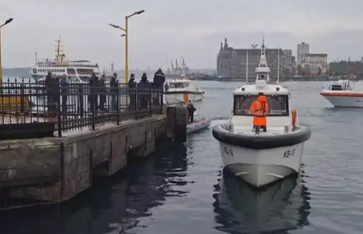 Bir iskelede kurtarma ekipleri ve polisler toplanmış. Denizde bir kurtarma botu ve birkaç tekne bulunuyor. Ekipler suya düşen kişileri kurtarmak için çalışıyor. Arka planda şehir manzarası ve binalar görünüyor.