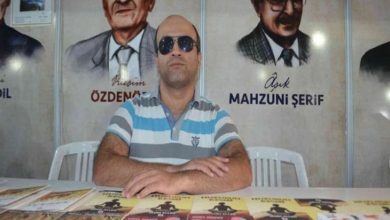 Görselde, masanın arkasında oturan bir erkek bulunuyor. Üzerinde mavi-beyaz çizgili bir tişört ve güneş gözlüğü var. Masada çeşitli kitaplar sergilenmiş. Arka planda duvarda ozan ve yazar portreleri ile isimleri yer alıyor.