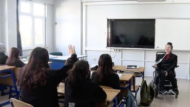 Görselde bir sınıf ortamı görülüyor. Öğretmen tekerlekli sandalyede, öğrencilerine doğru dönmüş şekilde ders anlatıyor. Ön sıradaki bir öğrenci el kaldırmış durumda. Öğretmen gülümserken, öğrenciler dikkatle onu dinliyor. Arka planda büyük bir akıllı tahta ve beyaz tahta yer alıyor.