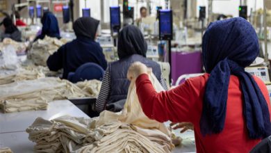 Görselde, bir tekstil atölyesinde çalışan kadın işçiler görülüyor. Başörtülü kadınlar, önlerinde yığılı kumaşlarla dikiş makinelerinde üretim yapıyor. Arka planda da benzer şekilde çalışan işçiler yer almakta. Çalışma ortamı yoğun ve üretim odaklı görünüyor.