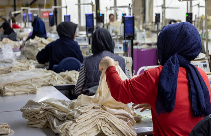 Görselde, bir tekstil atölyesinde çalışan kadın işçiler görülüyor. Başörtülü kadınlar, önlerinde yığılı kumaşlarla dikiş makinelerinde üretim yapıyor. Arka planda da benzer şekilde çalışan işçiler yer almakta. Çalışma ortamı yoğun ve üretim odaklı görünüyor.
