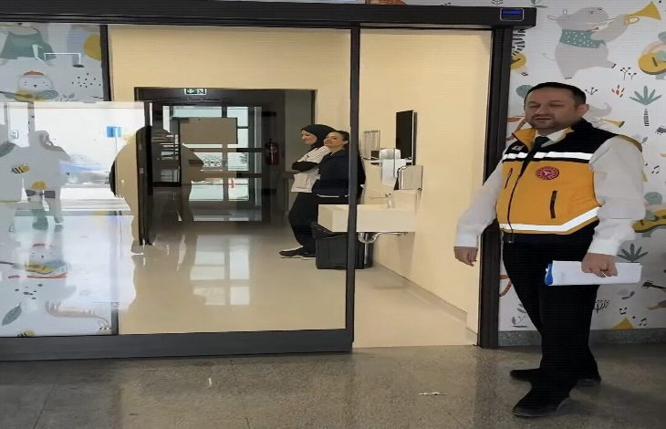 Görselde bir hastane ya da sağlık kurumunun girişinde duran bir görevli görülüyor. Görevli sarı-siyah yelek giymiş ve elinde bir belge tutuyor. Arka planda iki sağlık personeli koridorda ayakta duruyor. Duvarlarda çocuklara yönelik renkli desenler yer alıyor; ortam temiz ve aydınlık bir sağlık tesisi izlenimi veriyor.