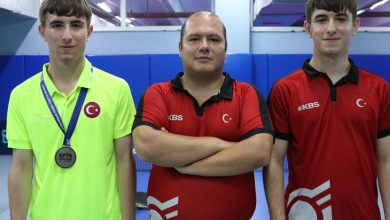 Görselde üç kişi spor salonunda poz veriyor. Ortadaki kişi kollarını göğsünde birleştirmiş, iki yanında duran gençler ise Türk bayraklı spor formaları giymiş. Soldaki gencin boynunda madalya asılı. Arka planda masa tenisi salonu ve masalar görülüyor.