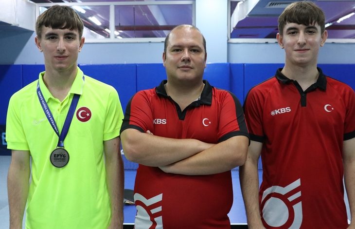 Görselde üç kişi spor salonunda poz veriyor. Ortadaki kişi kollarını göğsünde birleştirmiş, iki yanında duran gençler ise Türk bayraklı spor formaları giymiş. Soldaki gencin boynunda madalya asılı. Arka planda masa tenisi salonu ve masalar görülüyor.