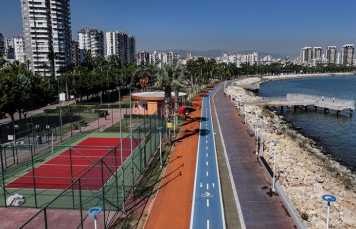 Mersin sahilinde modern bir yürüyüş ve bisiklet yolu yer almakta. Sol tarafta, yeşillikler içinde bir tenis kortu ve çevresinde palmiyelerle süslenmiş park alanı görülüyor. Sağ tarafta ise kayalık bir kıyı boyunca uzanan sahil yürüyüş yolu, denize doğru uzanan bir iskele ile tamamlanıyor. Arka planda ise yüksek apartmanlar ve şehir silueti, Akdeniz'in berrak maviliğiyle birleşerek ferahlatıcı bir atmosfer yaratıyor.