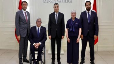 Görselde, beş kişi bir arada poz veriyor. Ortadaki kişi koyu renk takım elbise giymiş, yanında bir tekerlekli sandalyede oturan erkek, diğerleri ise ayakta duruyor. Arka planda “T.C. Mersin Valiliği” yazısı ve iki Türk bayrağı görülüyor. Fotoğraf resmî bir ziyaret veya toplantı sonrasında çekilmiş izlenimi veriyor.