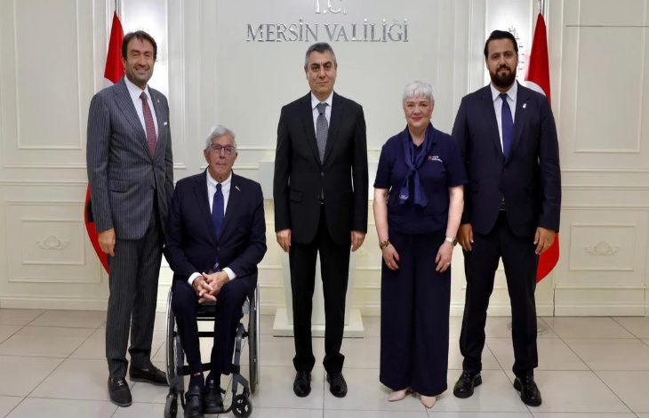 Görselde, beş kişi bir arada poz veriyor. Ortadaki kişi koyu renk takım elbise giymiş, yanında bir tekerlekli sandalyede oturan erkek, diğerleri ise ayakta duruyor. Arka planda “T.C. Mersin Valiliği” yazısı ve iki Türk bayrağı görülüyor. Fotoğraf resmî bir ziyaret veya toplantı sonrasında çekilmiş izlenimi veriyor.