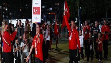 Görselde gece yapılan bir açılış töreninde Türk spor kafilesi kortej halinde yürüyor. Önde “Türkiye” yazılı bir pankart ve Türk bayrağı taşıyan kişiler var. Sporcular kırmızı eşofman giymiş, etraflarında alkışlayan izleyiciler bulunuyor. Arka planda açık hava etkinlik alanı ve ışıklandırmalar görülüyor.