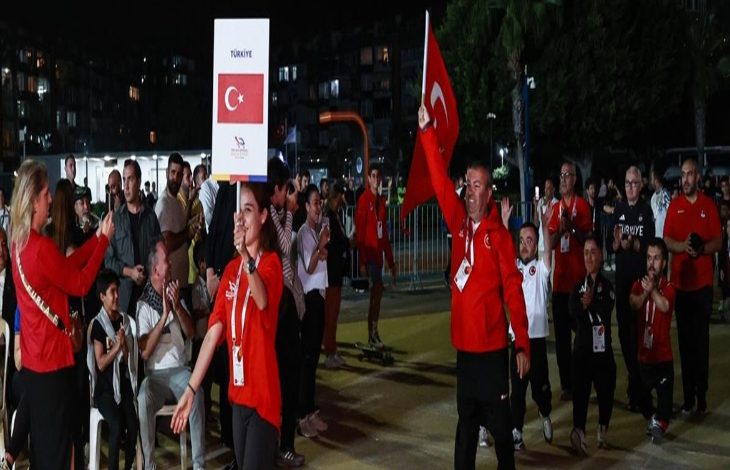 Görselde gece yapılan bir açılış töreninde Türk spor kafilesi kortej halinde yürüyor. Önde “Türkiye” yazılı bir pankart ve Türk bayrağı taşıyan kişiler var. Sporcular kırmızı eşofman giymiş, etraflarında alkışlayan izleyiciler bulunuyor. Arka planda açık hava etkinlik alanı ve ışıklandırmalar görülüyor.