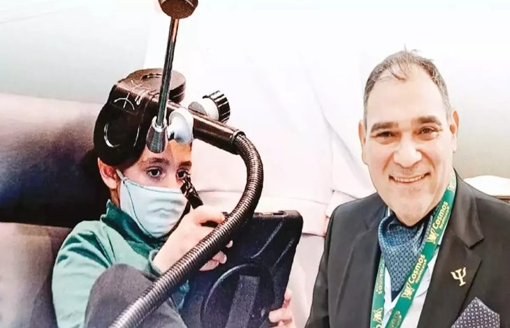 Görselde iki kişi yer alıyor. Solda, yüzünde maske olan bir çocuk başında özel bir cihazla tablet kullanıyor. Sağda ise takım elbiseli, boynunda kimlik kartı asılı gülümseyen bir yetişkin bulunuyor.