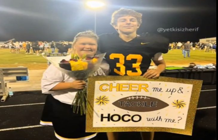 Bir futbol sahasında çekilmiş fotoğrafta, formasıyla bir erkek öğrenci ve çiçek tutan bir kız öğrenci yan yana duruyor. Erkek öğrenci elinde “Cheer me up & tackle HOCO with me?” yazılı süslenmiş bir pano tutuyor. Arkada gece ışıkları altında kalabalık bir grup görünüyor.