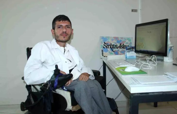 Psikolog Serkan Arslan, akülü sandalyede oturarak bilgisayar bulunan çalışma masasında poz veriyor.