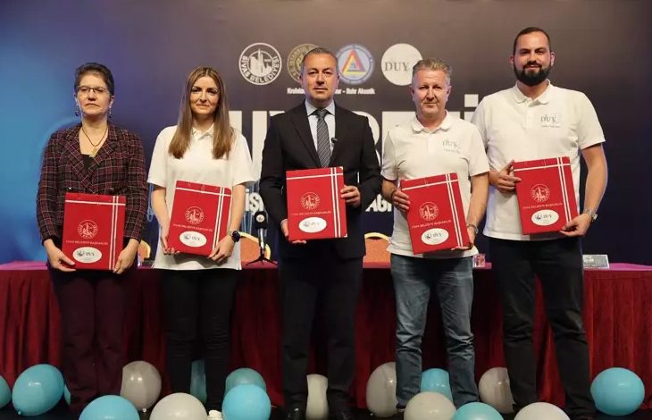 Görselde beş kişi yan yana duruyor. Ellerinde kırmızı renkli dosyalar tutuyorlar. Ortadaki kişi takım elbiseli, diğer dört kişi ise daha sade kıyafetli. Arka planda "DUY BENİ" yazısı ve bazı kurum logoları yer almakta. Ön tarafta ise süsleme amaçlı balonlar bulunuyor. Bu görüntü, resmi bir tören ya da proje tanıtımı kapsamında çekilmiş gibi görünüyor.