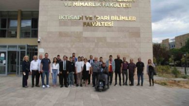 Görselde bir grup insan, üniversite binasının önünde hatıra fotoğrafı çektiriyor. Binanın duvarında “Güzel Sanatlar Tasarım ve Mimarlık Fakültesi” ile “İktisadi ve İdari Bilimler Fakültesi” yazıları yer alıyor. Ön sırada tekerlekli sandalyedeki bir kişi de bulunuyor. Katılımcılar resmi veya yarı resmi kıyafetlerle kameraya bakarak poz veriyor. Hava açık ve ortam kampüs atmosferinde.
