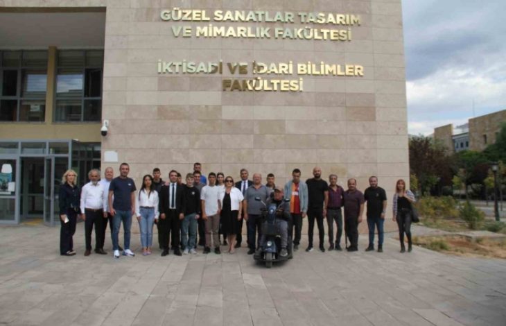 Görselde bir grup insan, üniversite binasının önünde hatıra fotoğrafı çektiriyor. Binanın duvarında “Güzel Sanatlar Tasarım ve Mimarlık Fakültesi” ile “İktisadi ve İdari Bilimler Fakültesi” yazıları yer alıyor. Ön sırada tekerlekli sandalyedeki bir kişi de bulunuyor. Katılımcılar resmi veya yarı resmi kıyafetlerle kameraya bakarak poz veriyor. Hava açık ve ortam kampüs atmosferinde.