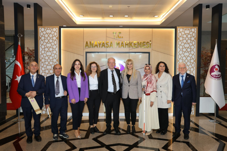 Türkiye Barolar Birliği Engelli Hakları Komisyonu üyeleri Anayasa Mahkemesi hizmet binası giriş alanında yan yana bulunuyor. Arka planda “T.C. ANAYASA MAHKEMESİ” yazısı ve kurumun amblemi dikkat çekerken Fotoğrafta, **Türkiye Cumhuriyeti Anayasa Mahkemesi** binasında çekilmiş resmi bir ziyaret anı yer alıyor. Arka planda “T.C. ANAYASA MAHKEMESİ” yazısı ve kurumun amblemi dikkat çekerken, sağ ve sol tarafta Türk bayrağı ile Anayasa Mahkemesi’nin kurumsal bayrağı yer almakta.