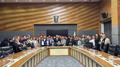 Görselde, Türkiye Büyük Millet Meclisi’ndeki bir toplantı salonunda kalabalık bir grup toplu fotoğraf için poz veriyor. Masanın etrafında kadın ve erkeklerden oluşan çok sayıda kişi bir arada. Arkada Türk bayrağı, bir başka bayrak ve Atatürk portresi bulunuyor. Ortam resmî ve toplantı atmosferinde.