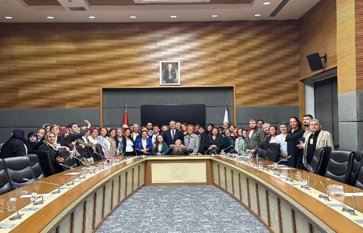 Görselde, Türkiye Büyük Millet Meclisi’ndeki bir toplantı salonunda kalabalık bir grup toplu fotoğraf için poz veriyor. Masanın etrafında kadın ve erkeklerden oluşan çok sayıda kişi bir arada. Arkada Türk bayrağı, bir başka bayrak ve Atatürk portresi bulunuyor. Ortam resmî ve toplantı atmosferinde.