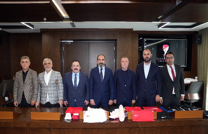 Görselde bir toplantı salonunda yedi erkek yan yana duruyor. Tam önlerinde kırmızı ve beyaz renkli spor kıyafetleri, ayakkabılar ve çeşitli ekipmanlar yer alıyor. Arka planda ahşap duvarlı bir ofis ortamı ve ekranda Türkiye Futbol Federasyonu’nun logosu görülüyor. Grup, resmi bir toplantı veya protokol buluşması için bir araya gelmiş izlenimi veriyor.