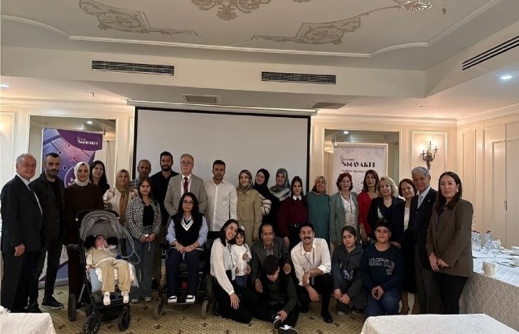 Görselde, bir toplantı salonunda kalabalık bir grup hatıra fotoğrafı çektiriyor. Katılımcılar farklı yaş ve gruplardan oluşuyor; aralarında tekerlekli sandalye kullanan ve bebek arabasıyla gelen kişiler de var. Arka planda bir projeksiyon perdesi ve etkinlik afişleri görülüyor. Ortam resmi ve samimi bir buluşma havasında.