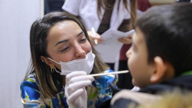 Maske ve eldiven takan bir sağlık çalışanı, küçük bir çocuğa pamuklu çubukla boğaz sürüntüsü testi yapıyor. Kadın, çocuğa gülümseyerek nazik bir şekilde yaklaşırken, arka planda başka sağlık personelleri görülüyor.