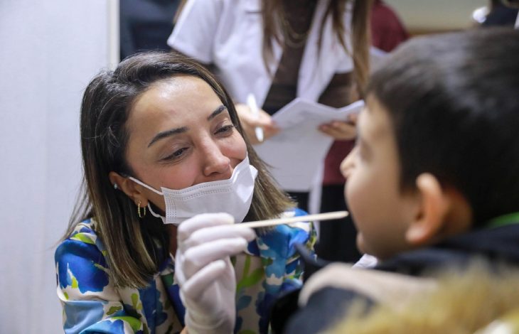 Maske ve eldiven takan bir sağlık çalışanı, küçük bir çocuğa pamuklu çubukla boğaz sürüntüsü testi yapıyor. Kadın, çocuğa gülümseyerek nazik bir şekilde yaklaşırken, arka planda başka sağlık personelleri görülüyor.