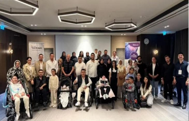 Bir toplantı salonunda, çoğunluğu ailelerden oluşan kalabalık bir grup birlikte poz veriyor. Ön sırada tekerlekli sandalyede ve bebek arabasında çocuklar bulunuyor.