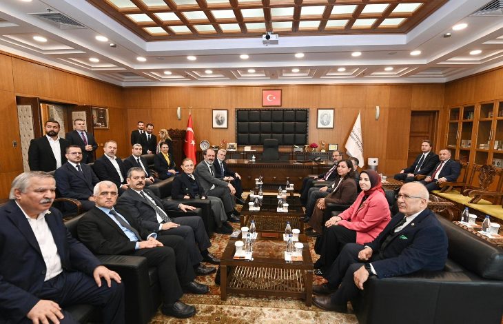 Geniş, resmi bir toplantı salonunda büyük bir masa etrafında oturan kalabalık bir heyet görülüyor. Takım elbiseli erkekler ve birkaç kadın, protokol düzeninde karşılıklı sıralanmış. Arka planda Türk bayrağı, makam koltuğu ve duvara asılı portreler yer alıyor. Atmosfer ciddi ve resmi.