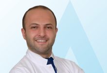 Beyaz önlük giymiş, gülümseyen bir erkek doktor Acıbadem International Hastanesi Pediatrik Ortopedi ve Travmatoloji Uzmanı Dr. Emre Sarıekiz, kameraya bakıyor. Arka plan açık mavi tonlarda sade bir tasarıma sahip.