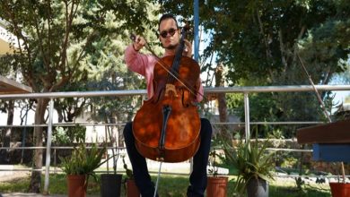 Açık alanda oturan bir erkek müzisyen, elinde yayla çello çalıyor. Arka planda ağaçlar ve saksı bitkileri görülüyor.