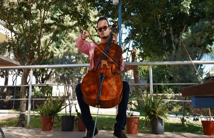 Açık alanda oturan bir erkek müzisyen, elinde yayla çello çalıyor. Arka planda ağaçlar ve saksı bitkileri görülüyor.