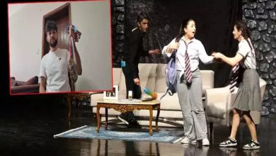 Bir sahne üzerinde üç genç oyuncu, okul kıyafetleriyle tiyatro performansı sergiliyor. Arkada kanepe ve masa bulunan bir salon dekoru yer alıyor. Görselin sol köşesinde ise bir erkek, elinde madalyalarla kameraya poz verirken görülüyor. Sahne enerjik ve duygusal bir atmosfer yansıtıyor.