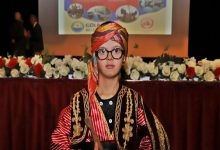 Görselde sahne önünde geleneksel bir seymen kıyafeti giymiş genç bir kişi bulunuyor. Üzerinde kırmızı-siyah tonlarda işlemeli, altın detaylı bir kostüm ve başında renkli bir sarık var. Elinde küçük bir davul tokmağı tutuyor. Arkada kırmızı çiçeklerle süslenmiş uzun bir masa ve üzerinde sunum yansıtılan bir sahne görünüyor. Etkinlik havası resme hâkim.