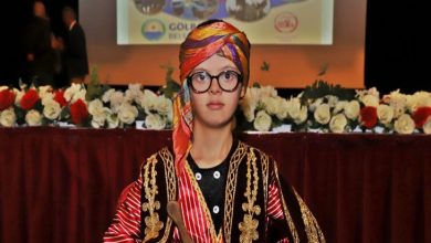 Görselde sahne önünde geleneksel bir seymen kıyafeti giymiş genç bir kişi bulunuyor. Üzerinde kırmızı-siyah tonlarda işlemeli, altın detaylı bir kostüm ve başında renkli bir sarık var. Elinde küçük bir davul tokmağı tutuyor. Arkada kırmızı çiçeklerle süslenmiş uzun bir masa ve üzerinde sunum yansıtılan bir sahne görünüyor. Etkinlik havası resme hâkim.