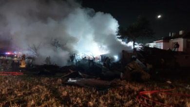 Görselde gece saatlerinde çıkan bir yangının ardından yoğun dumanın gökyüzüne yükseldiği bir alan görülüyor. İtfaiye ekipleri ve görevliler, alevlerin söndürüldüğü enkazın arasında çalışıyor. Önde yanmış malzemeler ve yıkıntılar yer alırken, arka planda itfaiye araçlarının ışıkları dumanın içinde parlıyor. Sahne, yangının büyük hasara yol açtığı ve müdahalenin hâlâ sürdüğü dramatik bir durumu yansıtıyor.