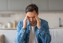 Bir koltukta oturan bir erkek, başının iki yanında şakaklarını tutmuş hâlde, yüzünü buruşturuyor. Sanki yoğun bir baş ağrısı yaşıyor ya da zihnini toparlamaya çalışıyor. Gündelik bir ev ortamında, loş ve sakin bir arka plan var. Atmosfer gergin ve rahatsızlık hissi veriyor.