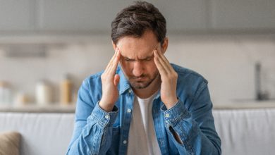 Bir koltukta oturan bir erkek, başının iki yanında şakaklarını tutmuş hâlde, yüzünü buruşturuyor. Sanki yoğun bir baş ağrısı yaşıyor ya da zihnini toparlamaya çalışıyor. Gündelik bir ev ortamında, loş ve sakin bir arka plan var. Atmosfer gergin ve rahatsızlık hissi veriyor.