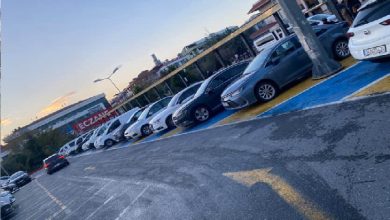 Görselde, yere mavi renkle işaretlenmiş engelli park yerlerinin tamamına yakınında araçların park ettiği bir otopark alanı görülüyor. Araçlar yan yana dizilmiş durumda ve engelli bireyler için ayrılan alanların dolu olduğu net biçimde anlaşılıyor. Arka planda binalar ve “Eczane” tabelası dikkat çekiyor. Sahne, engelli park yerlerinin sürekli meşgul olmasına dair yaşanan sorunu yansıtıyor.