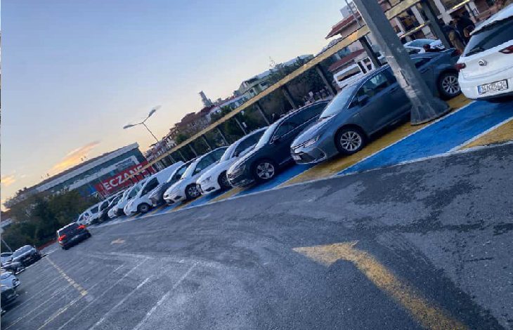 Görselde, yere mavi renkle işaretlenmiş engelli park yerlerinin tamamına yakınında araçların park ettiği bir otopark alanı görülüyor. Araçlar yan yana dizilmiş durumda ve engelli bireyler için ayrılan alanların dolu olduğu net biçimde anlaşılıyor. Arka planda binalar ve “Eczane” tabelası dikkat çekiyor. Sahne, engelli park yerlerinin sürekli meşgul olmasına dair yaşanan sorunu yansıtıyor.
