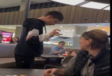 Görselde bir alışveriş merkezinin yemek alanında ayakta duran genç bir erkek çocuk görülüyor. Yan masadaki kadın ona doğru dönmüş durumda. Arka planda yemek yiyen başka kişiler de var. Ortam günlük, kalabalık ve hareketli bir food court havası taşıyor.