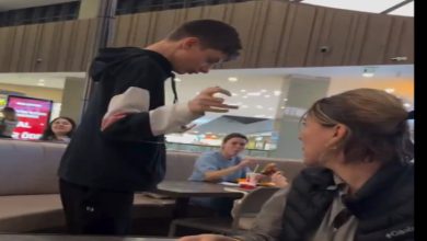 Görselde bir alışveriş merkezinin yemek alanında ayakta duran genç bir erkek çocuk görülüyor. Yan masadaki kadın ona doğru dönmüş durumda. Arka planda yemek yiyen başka kişiler de var. Ortam günlük, kalabalık ve hareketli bir food court havası taşıyor.