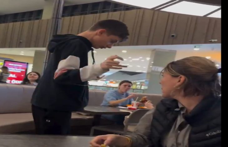 Görselde bir alışveriş merkezinin yemek alanında ayakta duran genç bir erkek çocuk görülüyor. Yan masadaki kadın ona doğru dönmüş durumda. Arka planda yemek yiyen başka kişiler de var. Ortam günlük, kalabalık ve hareketli bir food court havası taşıyor.