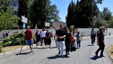 Görselde açık havada yürüyen bir grup insan görülüyor. Aralarında baston kullanan, koltuk değneğiyle yürüyen ve rehber eşliğinde hareket eden bireyler bulunuyor. Arka planda ağaçlar, binalar ve yön tabelaları yer almakta. Gün ışığı parlak, atmosfer canlı ve katılımcılar bir etkinlik ya da yürüyüş kapsamında bir araya gelmiş gibi görünüyor.