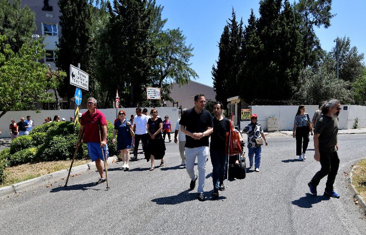 Görselde açık havada yürüyen bir grup insan görülüyor. Aralarında baston kullanan, koltuk değneğiyle yürüyen ve rehber eşliğinde hareket eden bireyler bulunuyor. Arka planda ağaçlar, binalar ve yön tabelaları yer almakta. Gün ışığı parlak, atmosfer canlı ve katılımcılar bir etkinlik ya da yürüyüş kapsamında bir araya gelmiş gibi görünüyor.