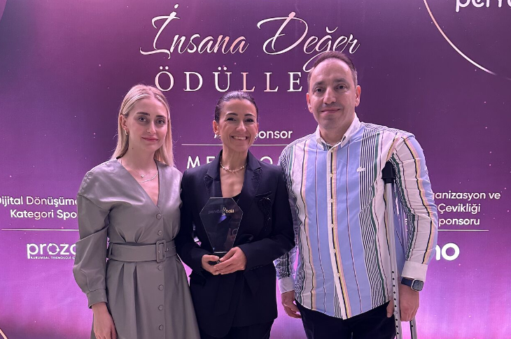 Üç kişinin yan yana durduğu bir ödül töreni pozu görünüyor. Ortadaki kişi elinde cam bir ödül tutuyor ve gülümseyerek kameraya bakıyor. Solunda gri elbiseli bir kadın, sağında ise çizgili gömlekli bir erkek yer alıyor. Arka planda mor tonlarda bir sahne dekoru ve “İnsana Değer Ödülleri” yazısı seçiliyor. Atmosfer tam bir kutlama ve gurur anı.